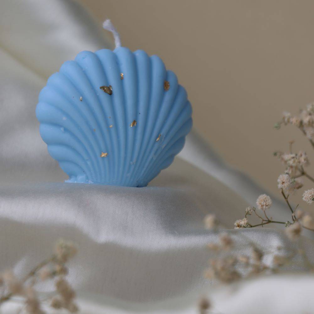 Blue shell-shaped candle with gold flakes, set on silky fabric with delicate floral accents. A nature-inspired decor piece for outdoor ambiance.