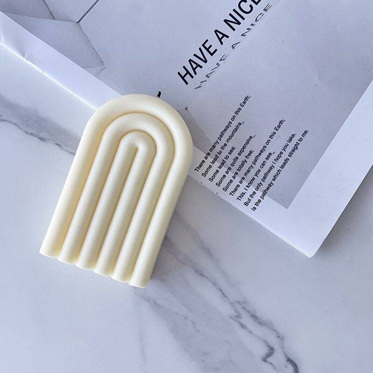 Minimalist rainbow-shaped candle in white, placed on a marble surface with a magazine. Elegant home fragrance decor for modern interiors.